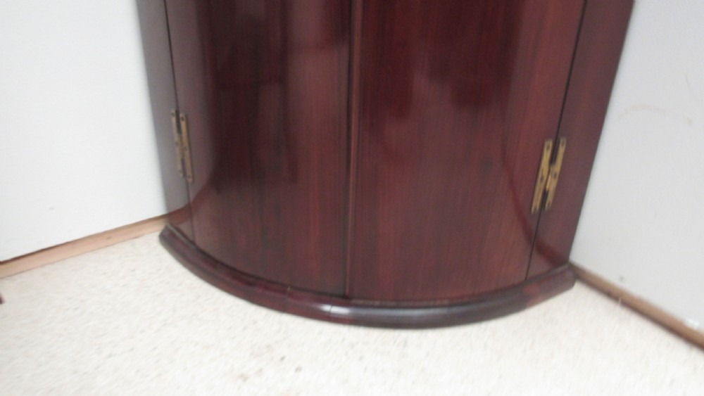 An antique Georgian bowfront corner cupboard crafted from dark mahogany with twin doors, brass H-hinges, and decorative escutcheons. View V