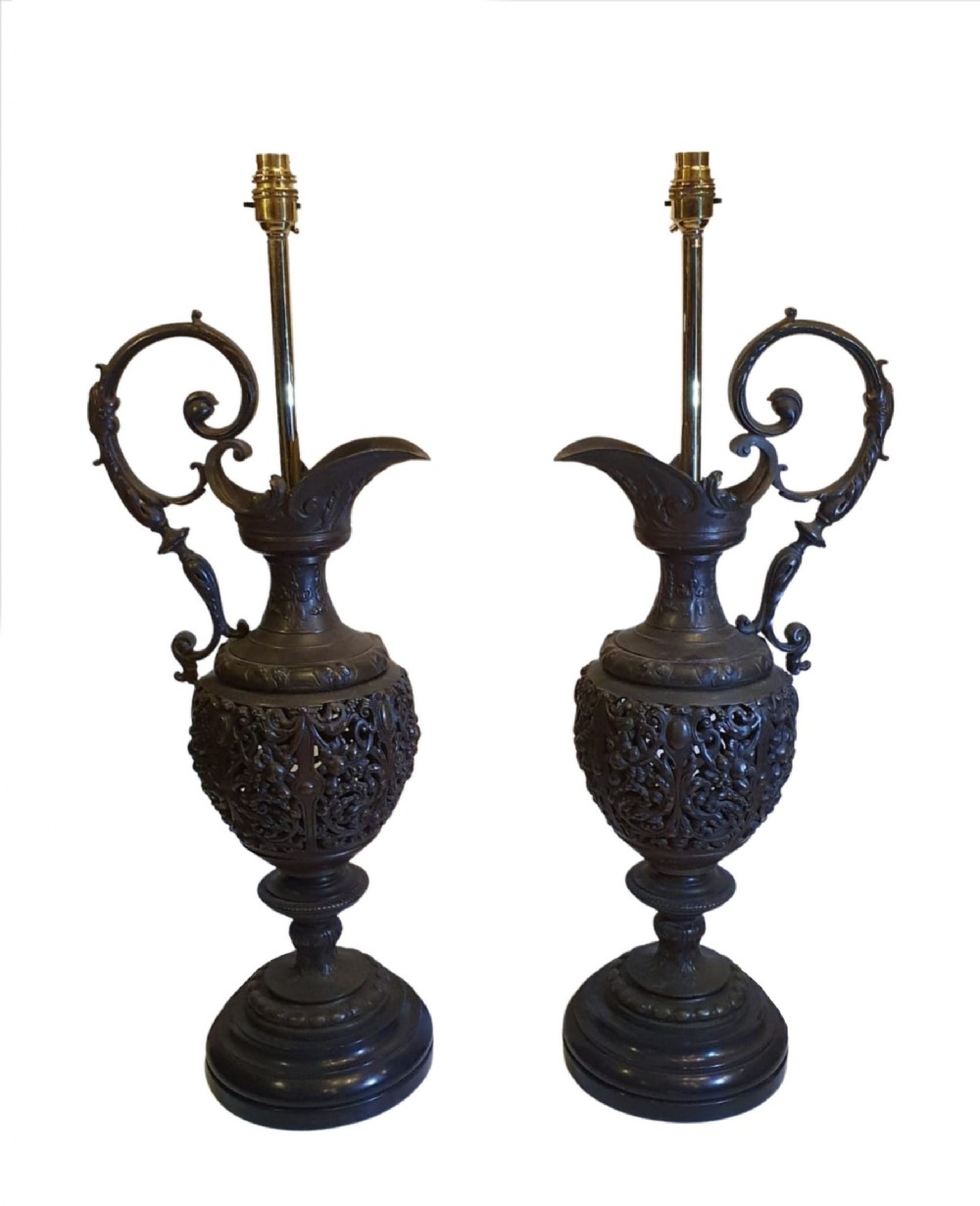 A pair of dark patinated bronze ewers featuring openwork scroll detail, high handles, and spout tops, converted into table lamps with brass electrical fittings.