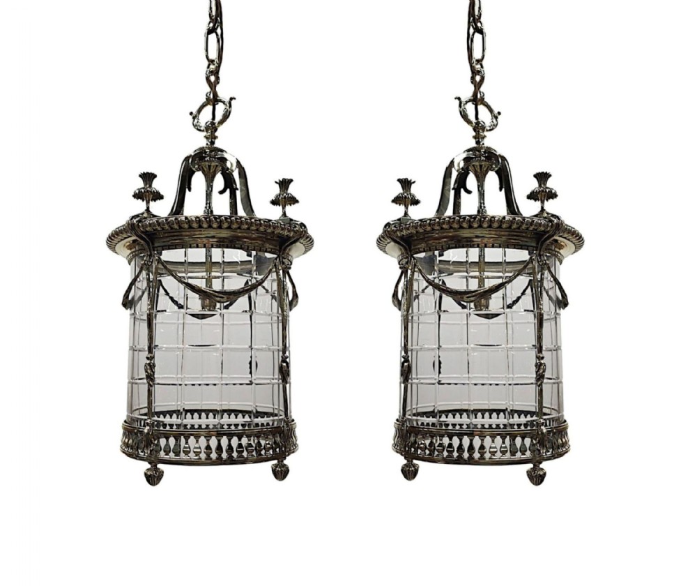 A pair of early 20th-century cylindrical glass hall lanterns featuring ornate silvered brass frames, hanging chains, draped metal swags, and decorative urn finials.