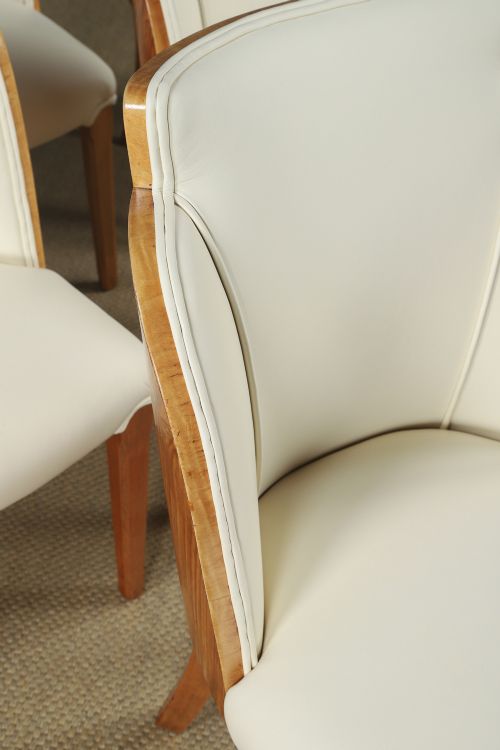 fig 3. An English Art Deco dining suite featuring a rectangular satinwood and maple dining table with a glass top and six cream leather upholstered chairs with curved veneered backs.