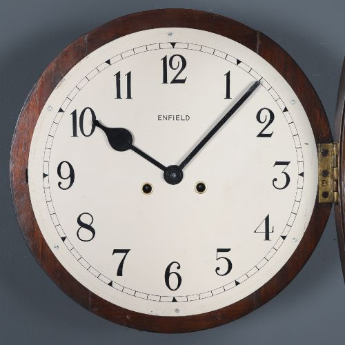 fig 3. A circular mahogany wall clock with a cream dial, black Arabic numerals, black metal hands, two brass winding holes, and the Enfield maker name printed in the centre.