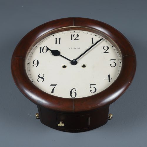 fig 7. A circular mahogany wall clock with a cream dial, black Arabic numerals, black metal hands, two brass winding holes, and the Enfield maker name printed in the centre.