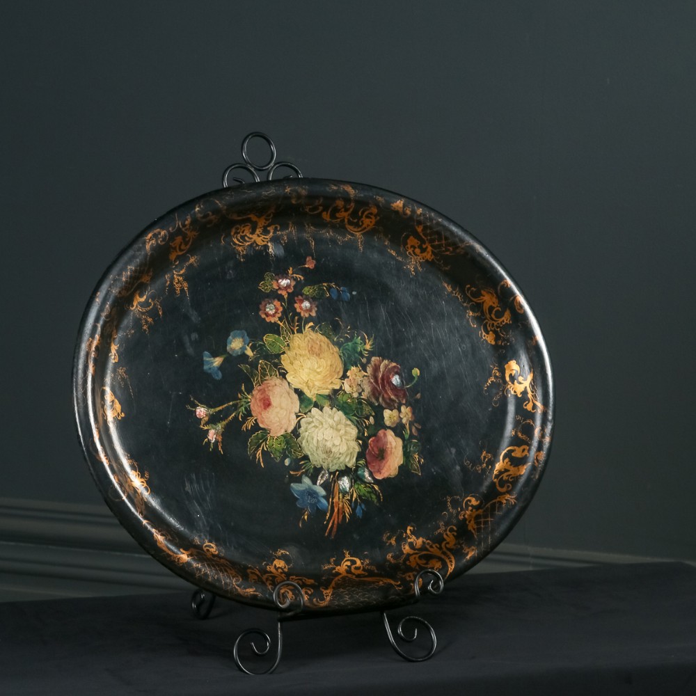 An oval ebonised papier-mache serving tray featuring a hand-painted floral bouquet centrepiece surrounded by gilt scrollwork detailing along the rim.