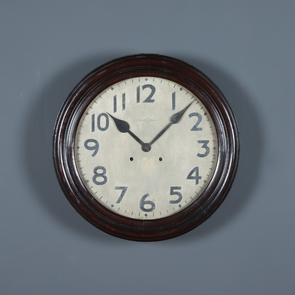 A large circular antique wall clock featuring a dark mahogany wooden surround, an off-white dial with bold black Arabic numerals, and twin winding keyholes.