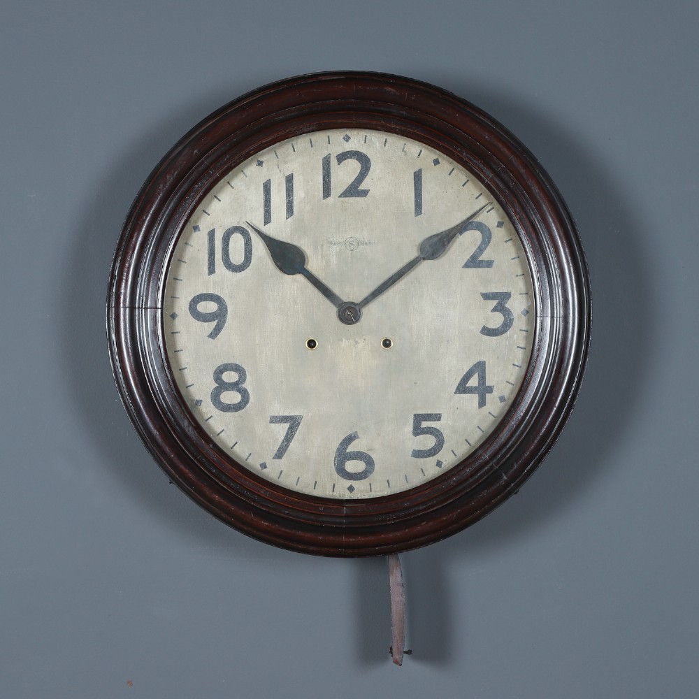 A large circular antique wall clock featuring a dark mahogany wooden surround, an off-white dial with bold black Arabic numerals, and twin winding keyholes. View V