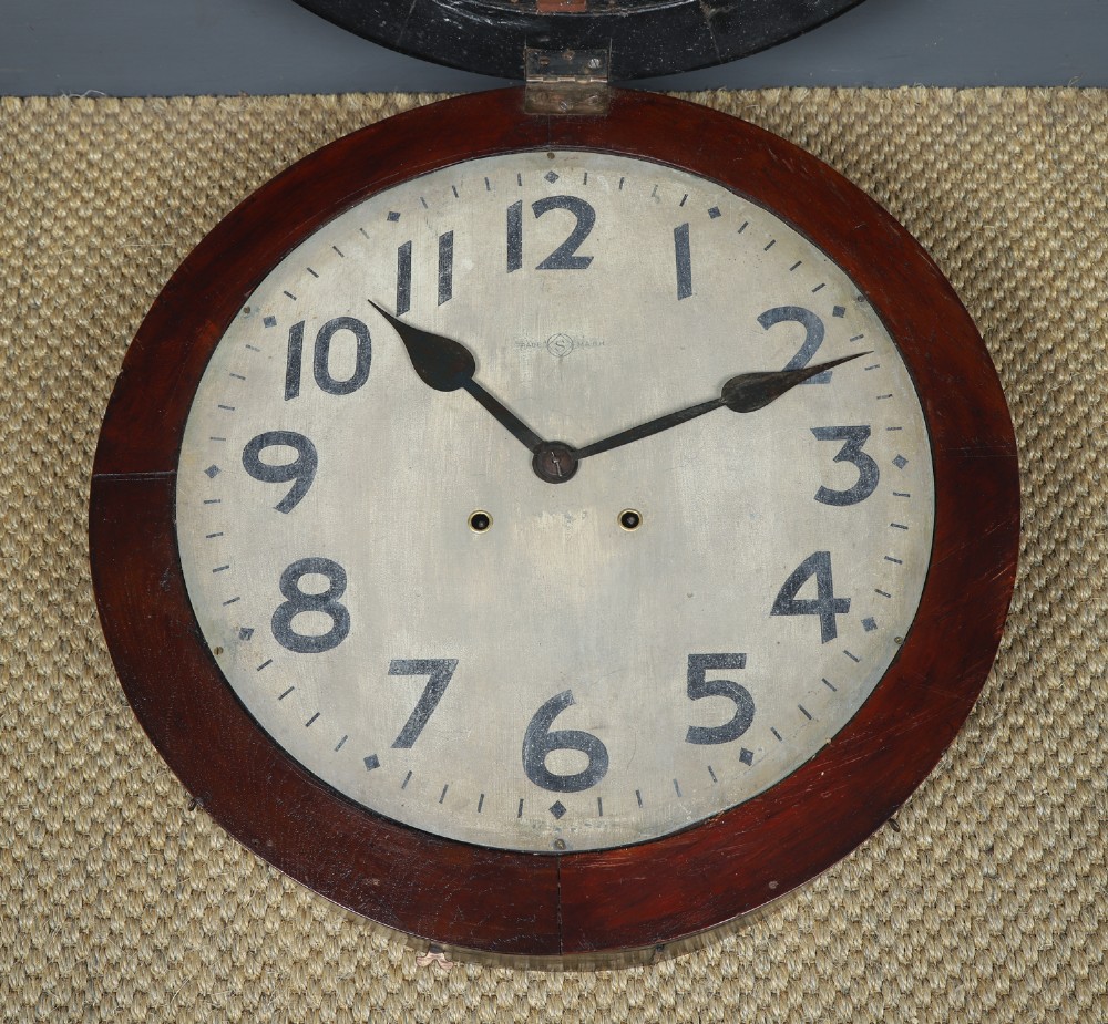 A large circular antique wall clock featuring a dark mahogany wooden surround, an off-white dial with bold black Arabic numerals, and twin winding keyholes. View VII