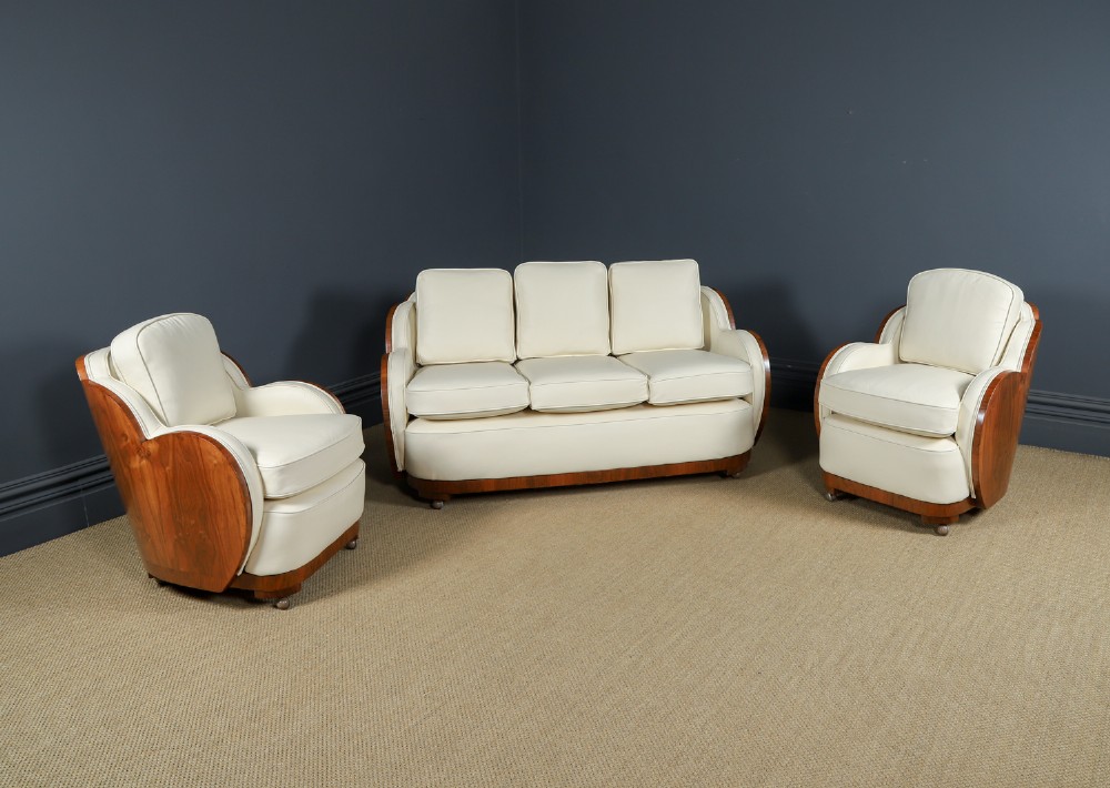 An antique Art Deco three-piece lounge suite consisting of a three-seater sofa and two matching armchairs upholstered in cream leather with curved walnut side panels.