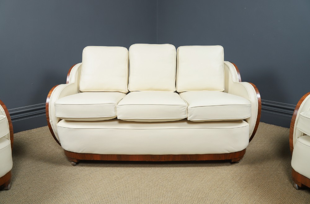 An antique Art Deco three-piece lounge suite consisting of a three-seater sofa and two matching armchairs upholstered in cream leather with curved walnut side panels. View V