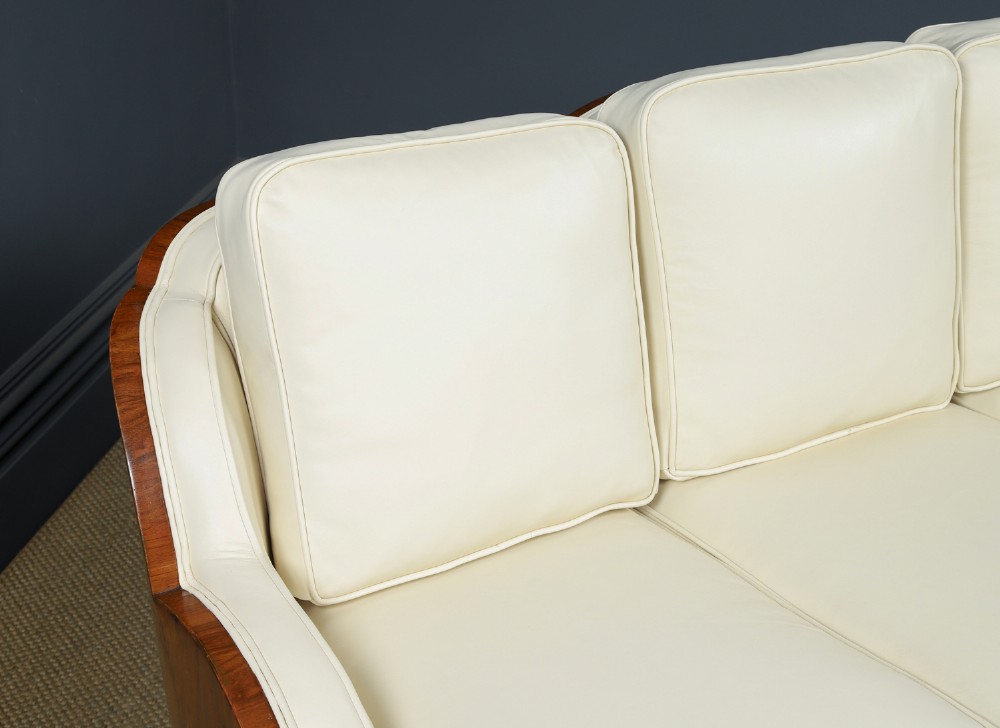 An antique Art Deco three-piece lounge suite consisting of a three-seater sofa and two matching armchairs upholstered in cream leather with curved walnut side panels. View VI