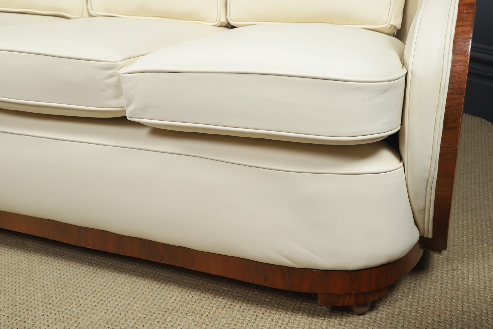 An antique Art Deco three-piece lounge suite consisting of a three-seater sofa and two matching armchairs upholstered in cream leather with curved walnut side panels. View IX