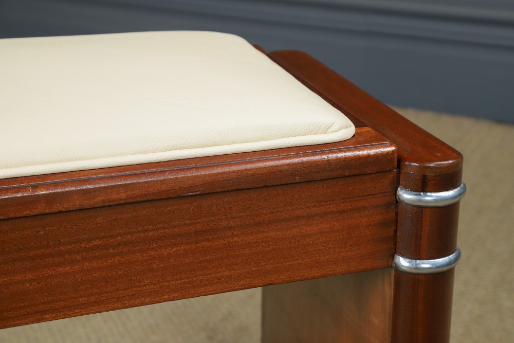 An Art Deco rectangular walnut stool featuring slab legs with metal band accents and a cream leather padded seat. View IV