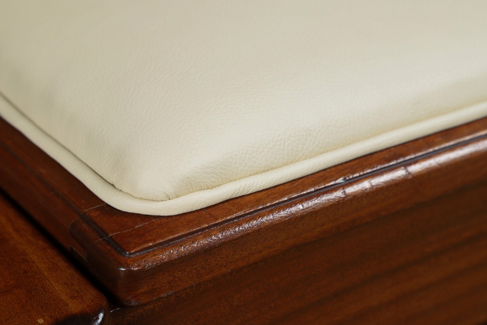 An Art Deco rectangular walnut stool featuring slab legs with metal band accents and a cream leather padded seat. View VI