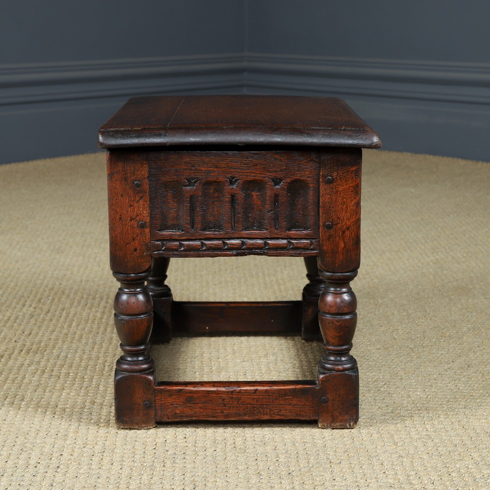 A small antique dark oak box stool featuring a hinged top lid, hand-carved frieze panels, turned legs, and square perimeter stretchers. View XII