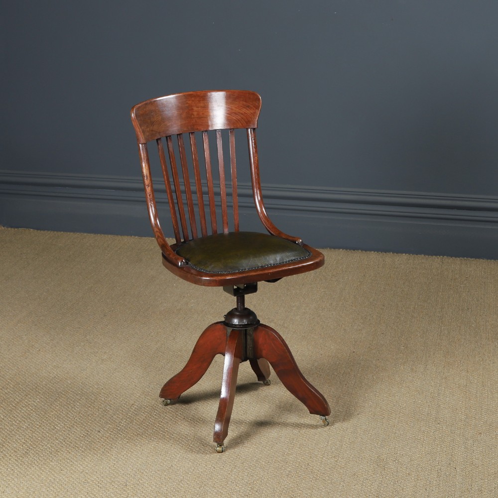 An antique George V stained beech revolving office chair featuring a slatted backrest, green leather seat, and splayed legs on brass castors.