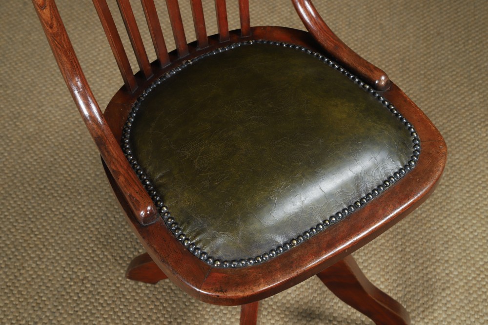 An antique George V stained beech revolving office chair featuring a slatted backrest, green leather seat, and splayed legs on brass castors. View IV