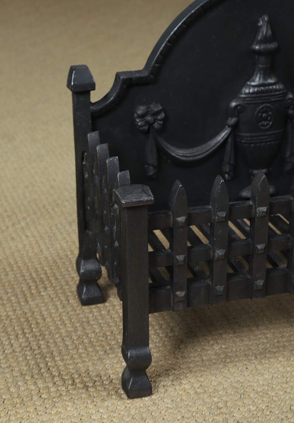 A black cast iron open fire grate featuring an arched backplate decorated with an urn relief and a palisade front basket resting on four square legs. View V