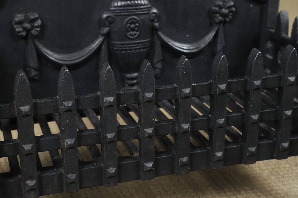 A black cast iron open fire grate featuring an arched backplate decorated with an urn relief and a palisade front basket resting on four square legs. View VII
