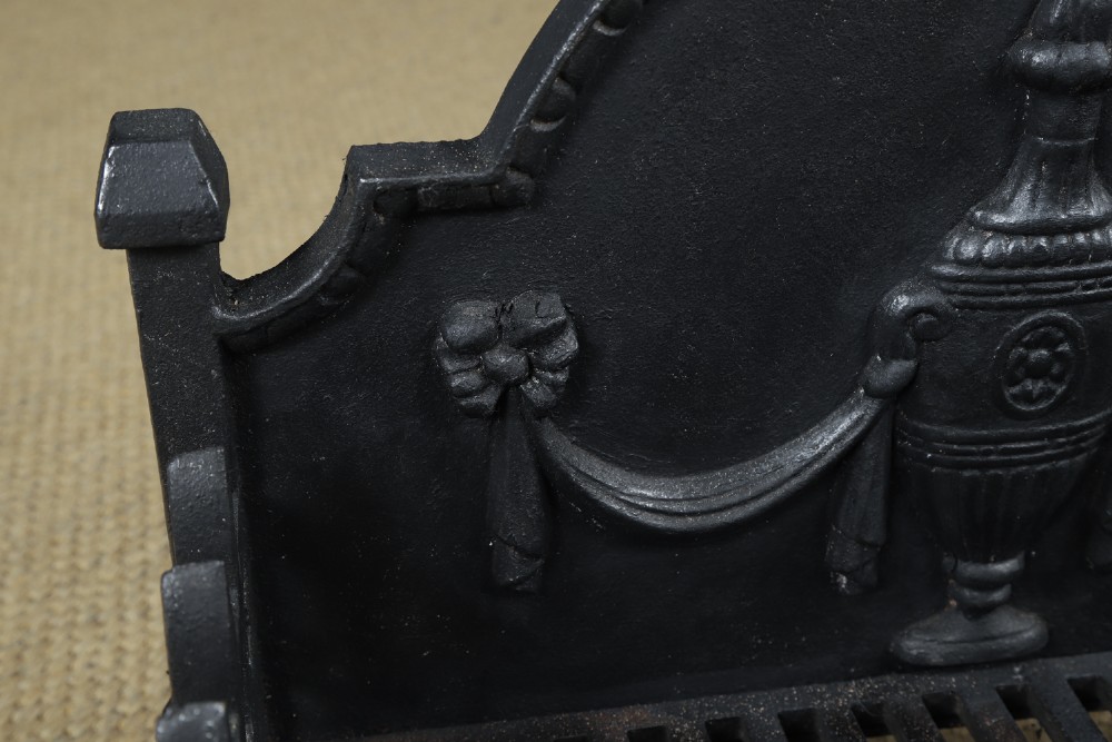A black cast iron open fire grate featuring an arched backplate decorated with an urn relief and a palisade front basket resting on four square legs. View IX