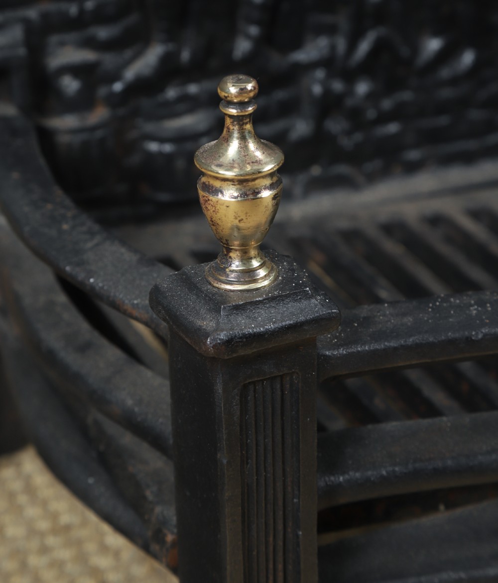 A black cast iron fire grate featuring an arched pictorial backplate, curved front bars, and brass urn finials on the front posts. View VI