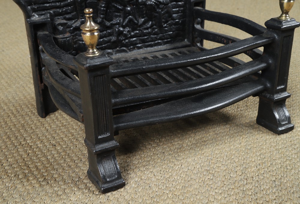 A black cast iron fire grate featuring an arched pictorial backplate, curved front bars, and brass urn finials on the front posts. View VII