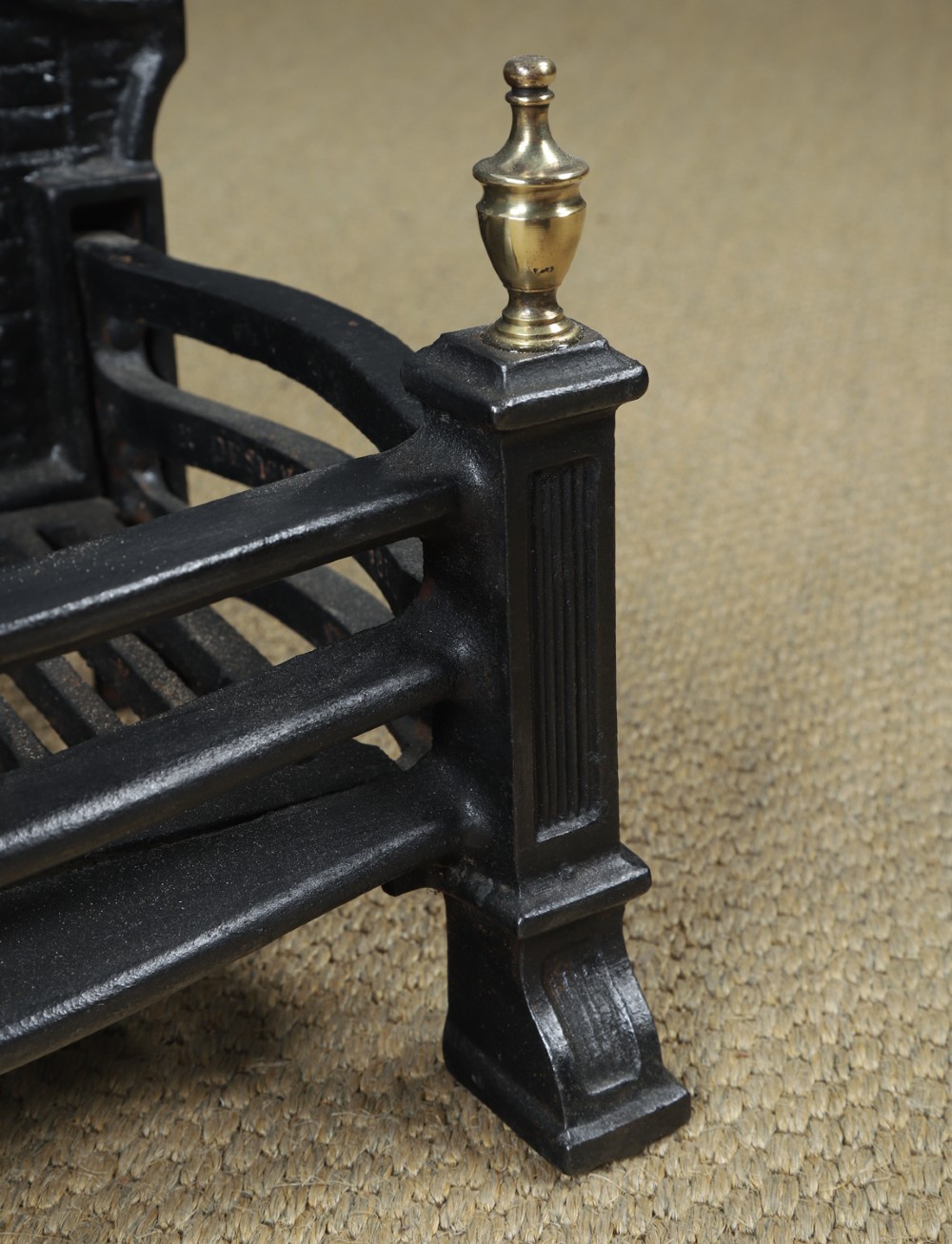 A black cast iron fire grate featuring an arched pictorial backplate, curved front bars, and brass urn finials on the front posts. View IX