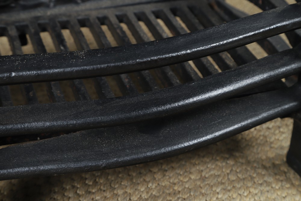 A black cast iron fire grate featuring an arched pictorial backplate, curved front bars, and brass urn finials on the front posts. View X
