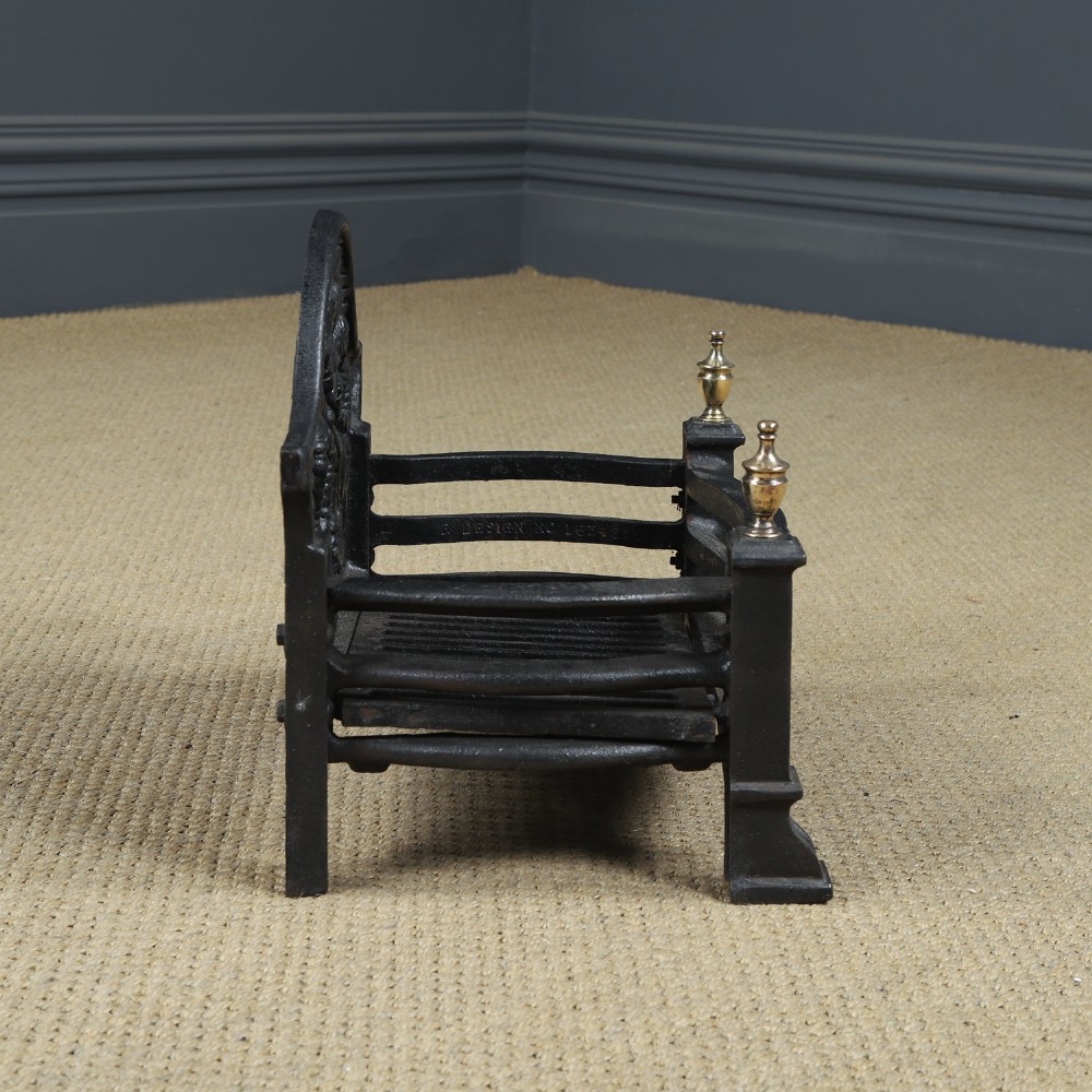 A black cast iron fire grate featuring an arched pictorial backplate, curved front bars, and brass urn finials on the front posts. View XI