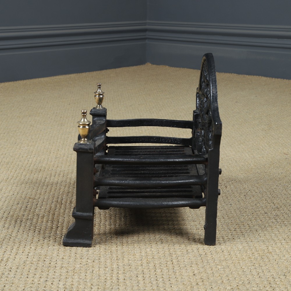 A black cast iron fire grate featuring an arched pictorial backplate, curved front bars, and brass urn finials on the front posts. View XII