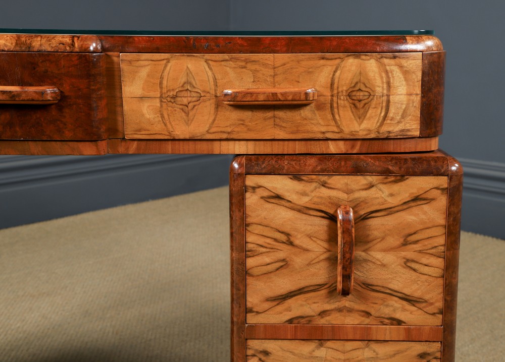 An Art Deco walnut and burr elm pedestal desk featuring stepped side pedestals, bookmatched wood veneers, wooden handles, and a fitted glass top. View X