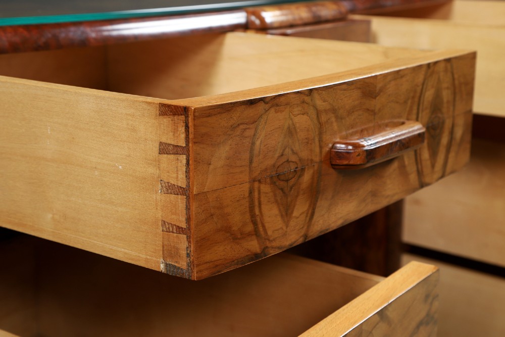 An Art Deco walnut and burr elm pedestal desk featuring stepped side pedestals, bookmatched wood veneers, wooden handles, and a fitted glass top. View XII