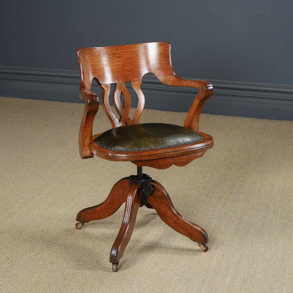 An Edwardian oak revolving desk armchair featuring a carved pierced back splat, green leather upholstered seat with close studding, and a four-point swivel base on brass castors.