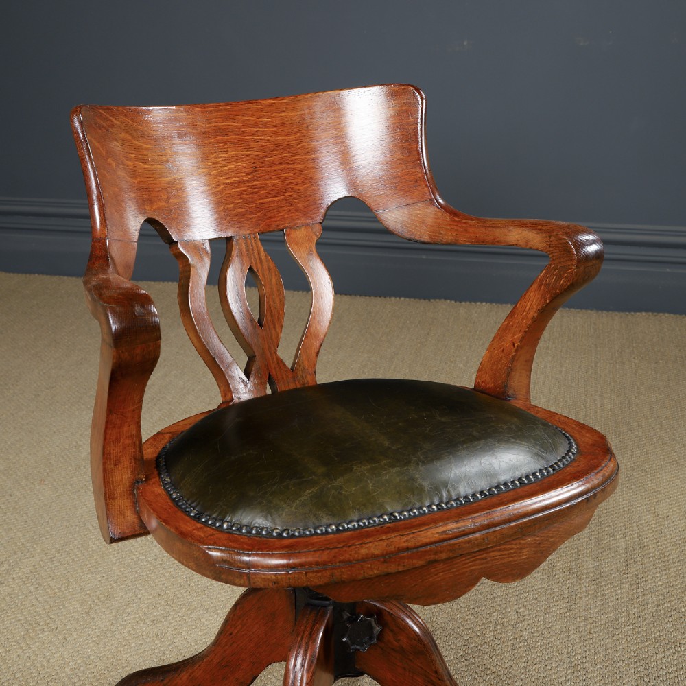 An Edwardian oak revolving desk armchair featuring a carved pierced back splat, green leather upholstered seat with close studding, and a four-point swivel base on brass castors. View II