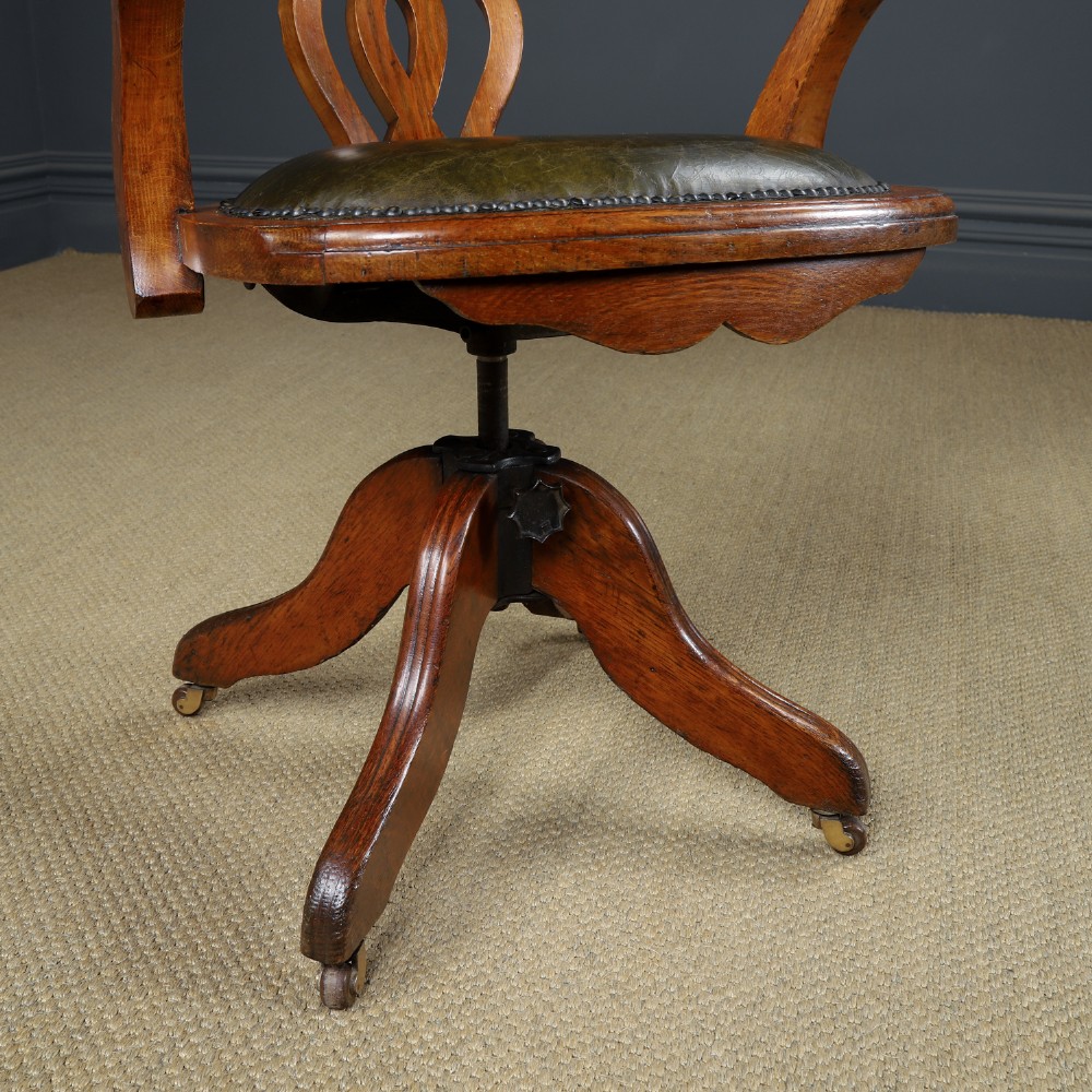 An Edwardian oak revolving desk armchair featuring a carved pierced back splat, green leather upholstered seat with close studding, and a four-point swivel base on brass castors. View III