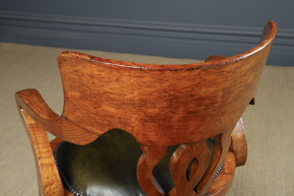 An Edwardian oak revolving desk armchair featuring a carved pierced back splat, green leather upholstered seat with close studding, and a four-point swivel base on brass castors. View XI