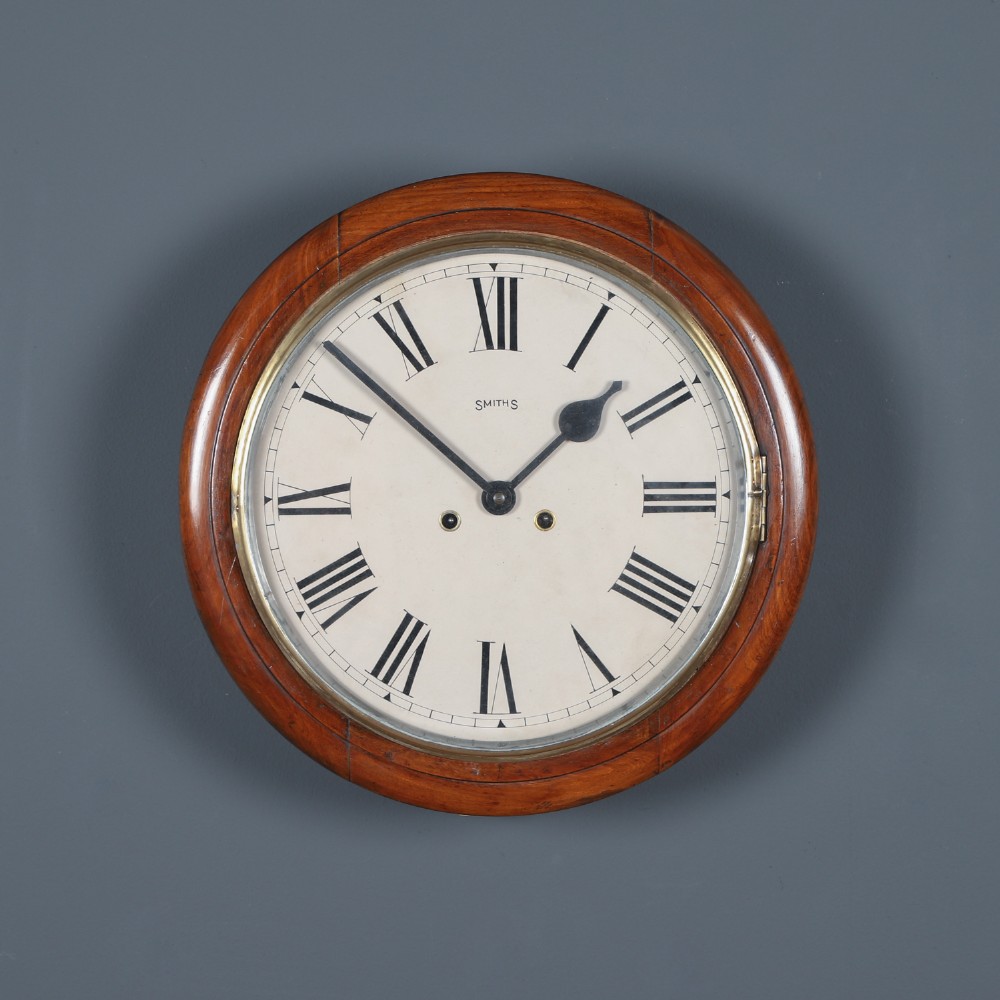 A circular Smiths antique wall clock with a turned mahogany wood surround, off-white dial with black Roman numerals, brass hinged bezel, and winding holes.