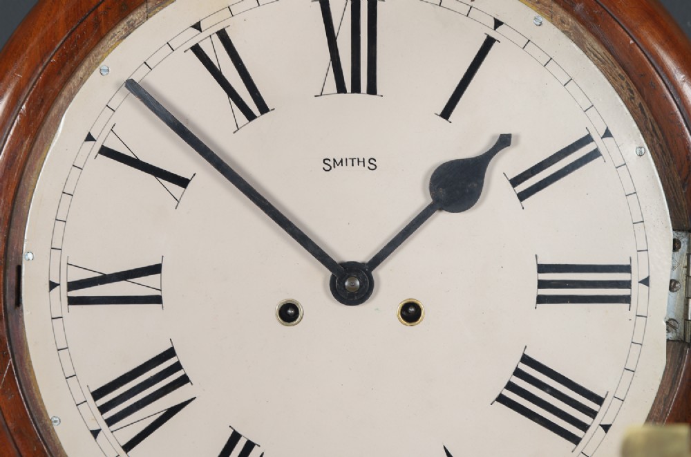 A circular Smiths antique wall clock with a turned mahogany wood surround, off-white dial with black Roman numerals, brass hinged bezel, and winding holes. View III