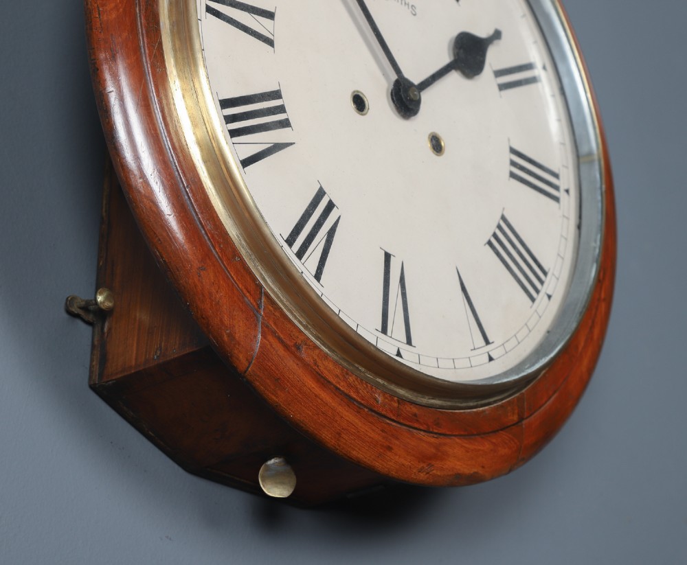 A circular Smiths antique wall clock with a turned mahogany wood surround, off-white dial with black Roman numerals, brass hinged bezel, and winding holes. View VI