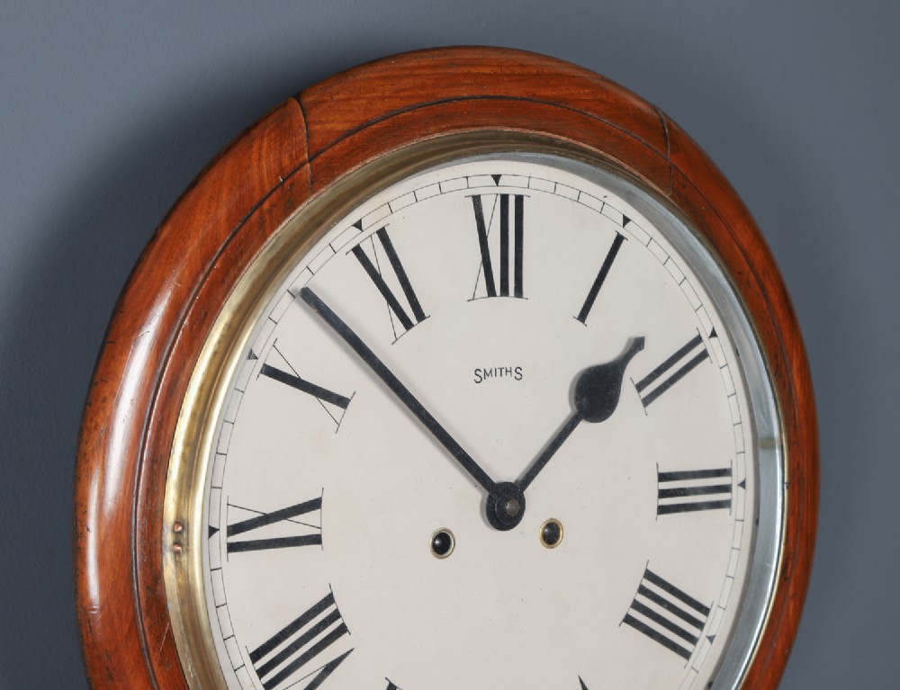 A circular Smiths antique wall clock with a turned mahogany wood surround, off-white dial with black Roman numerals, brass hinged bezel, and winding holes. View VII