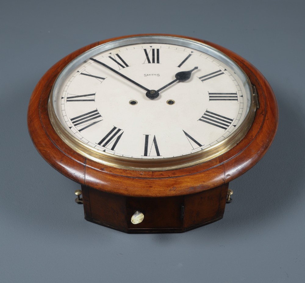 A circular Smiths antique wall clock with a turned mahogany wood surround, off-white dial with black Roman numerals, brass hinged bezel, and winding holes. View IX