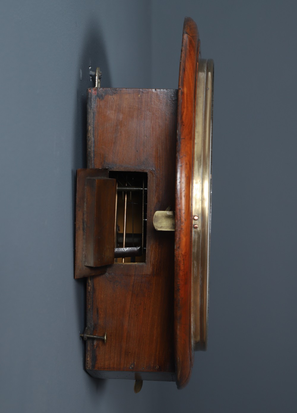 A circular Smiths antique wall clock with a turned mahogany wood surround, off-white dial with black Roman numerals, brass hinged bezel, and winding holes. View XI