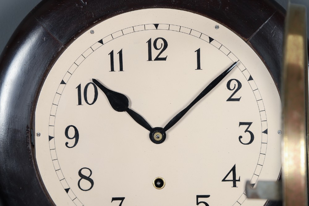 A circular antique dark mahogany wall clock featuring a white dial with black Arabic numerals and a brass bezel. View II
