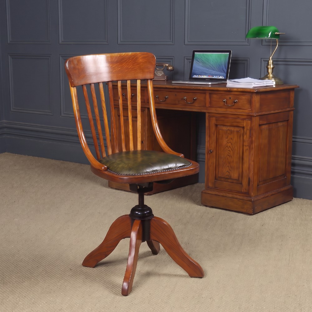 A George V period golden oak revolving desk chair featuring a curved spindle backrest, quadruped base, and a padded green leather seat secured with brass studs.
