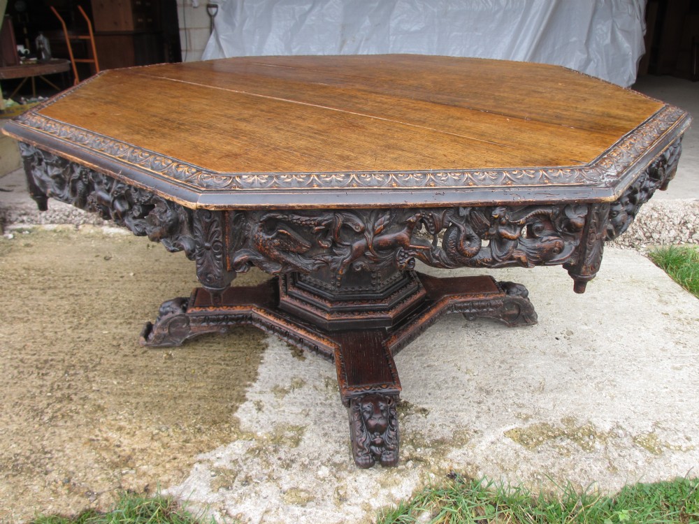 Superb 19th Century carved oak octagonal tilt top centre dining table with carved edge and fret carved mythical creature frieze with drop finials, carved studded tapering column, carved quadruple base set on castors