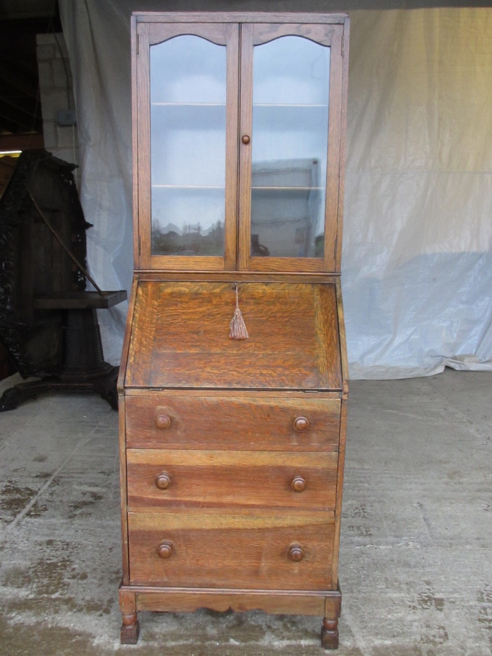 A narrow Edwardian oak bureau bookcase with twin glazed doors on the upper section above a fall-front writing desk and three drawers with wooden bun handles.