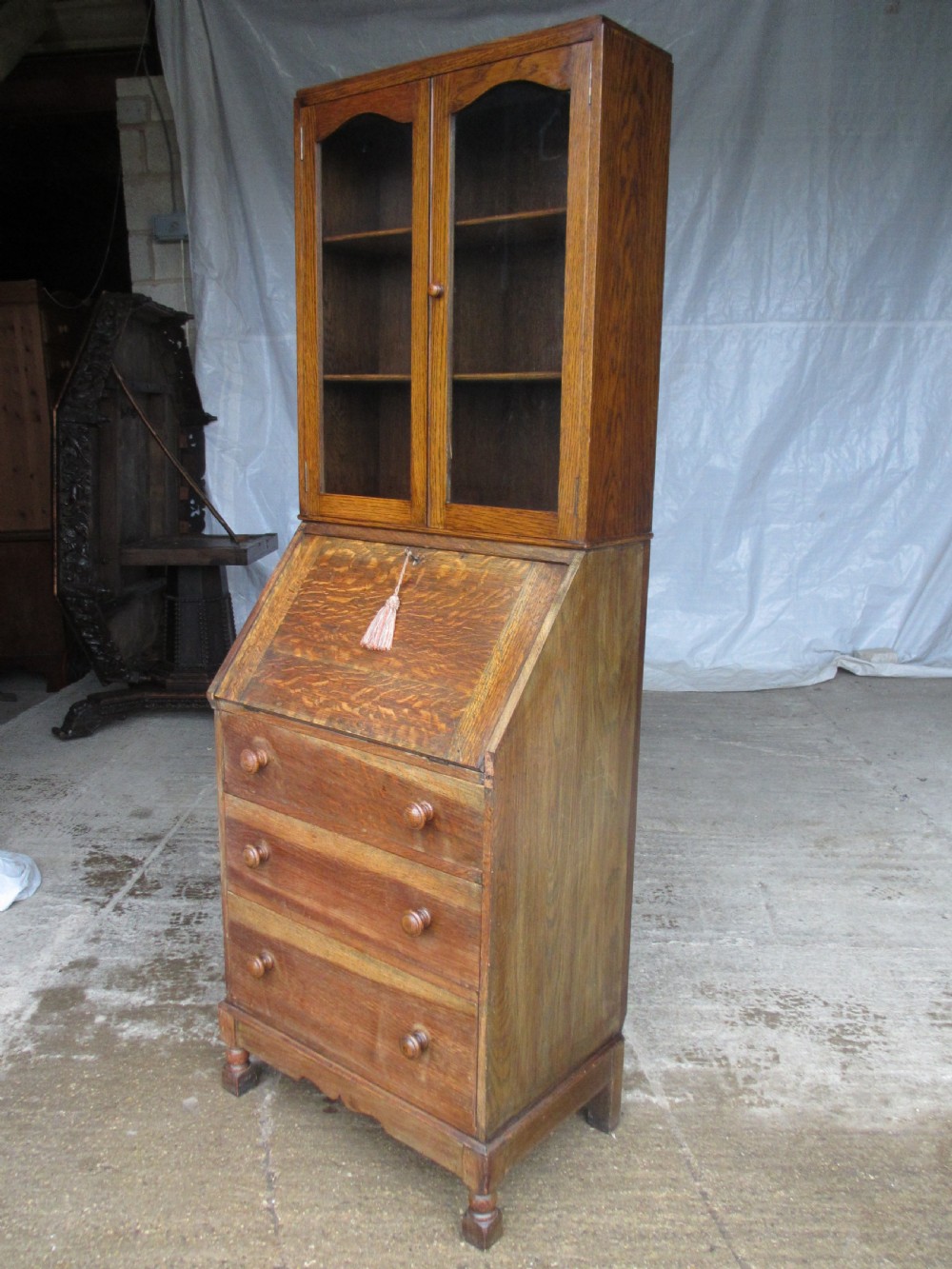 A narrow Edwardian oak bureau bookcase with twin glazed doors on the upper section above a fall-front writing desk and three drawers with wooden bun handles. View II
