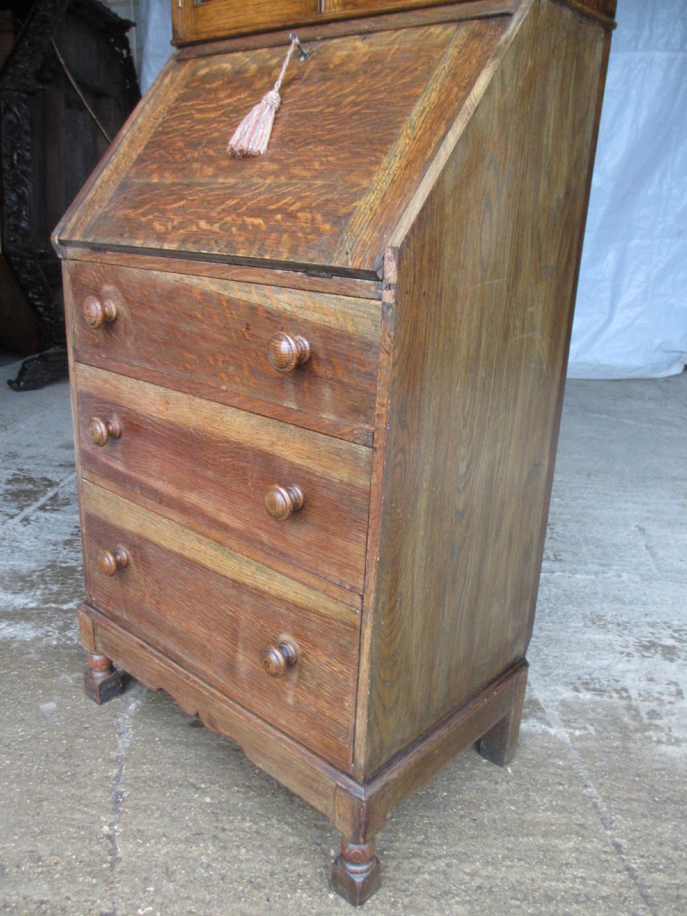 A narrow Edwardian oak bureau bookcase with twin glazed doors on the upper section above a fall-front writing desk and three drawers with wooden bun handles. View IV