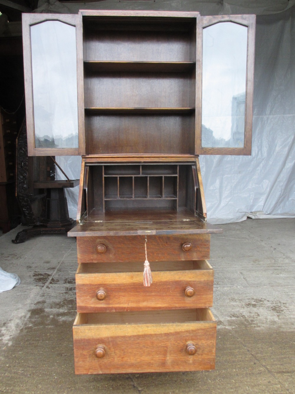 A narrow Edwardian oak bureau bookcase with twin glazed doors on the upper section above a fall-front writing desk and three drawers with wooden bun handles. View XII