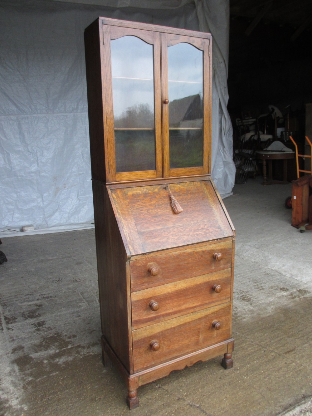 A narrow Edwardian oak bureau bookcase with twin glazed doors on the upper section above a fall-front writing desk and three drawers with wooden bun handles. View XIII