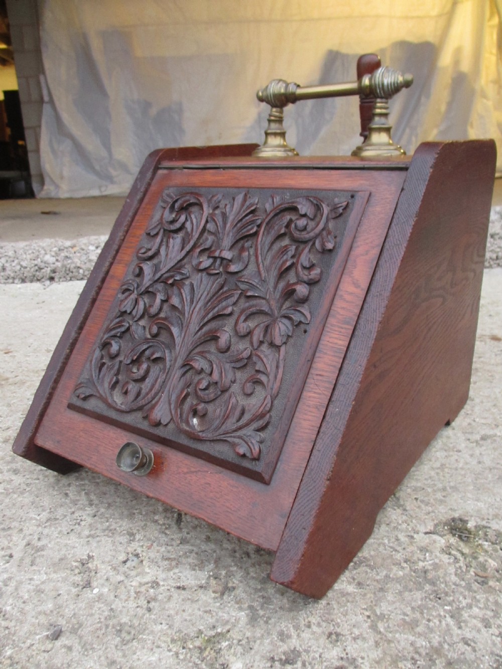 A Victorian dark oak purdonium coal box with a slanted front featuring a relief-carved acanthus leaf panel and a top brass handle.