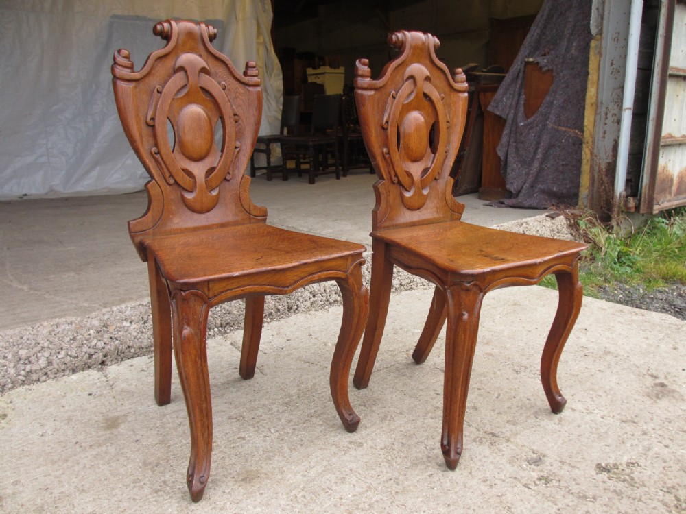 A pair of Victorian carved golden oak hall chairs featuring openwork shield backs with central cartouches, solid wooden seats, and cabriole front legs.